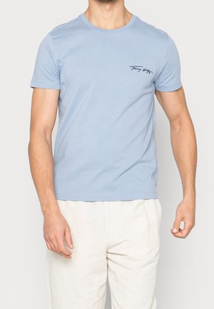 Mand iført lyseblå t-shirt med korte ærmer med lille "Tommy Hilfiger" tekst på venstre bryst og off-white bukser mod ensfarvet baggrund.