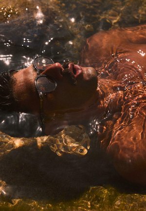 Bajszos férfi napszemüvegben, aki hanyatt fekve lebeg sekély vízben, napsütés tükröződik a víz felszínén.