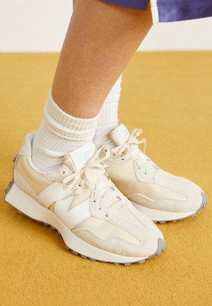 Osoba nosząca kremowe sneakersy New Balance z białymi sznurówkami i białe prążkowane skarpetki stojąca na żółtej podłodze.