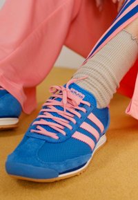 Sapatilhas azuis com detalhes cor-de-rosa, materiais em malha e camurça, apresentando três riscas e atacadores cor-de-rosa. Usadas com meias cinzentas caneladas.
