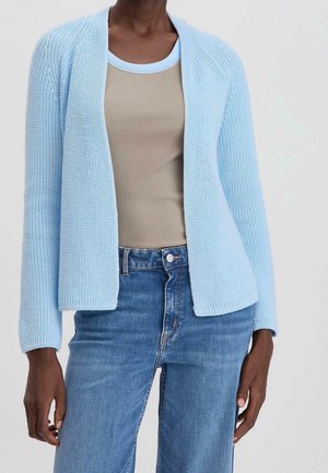 Vrouw draagt een lichtblauwe opengewerkte cardigan over een beige geribd tanktopje en een high-waisted blauwe spijkerbroek tegen een effen achtergrond.
