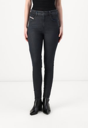 Femme portant un jean skinny noir, un haut sans manches noir et des bottines à talons noires, debout sur un fond blanc.