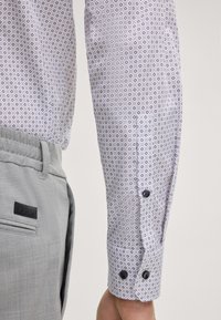 Chemise blanche avec de petits motifs circulaires répétés en bleu et rouge. Pantalon gris en tissu texturé avec une étiquette noire sur la ceinture.