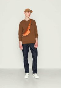 Sweatshirt castanho, jeans escuros e ténis brancos. Modelo a usar um boné castanho e uma bolsa cruzada laranja com fecho de zíper.