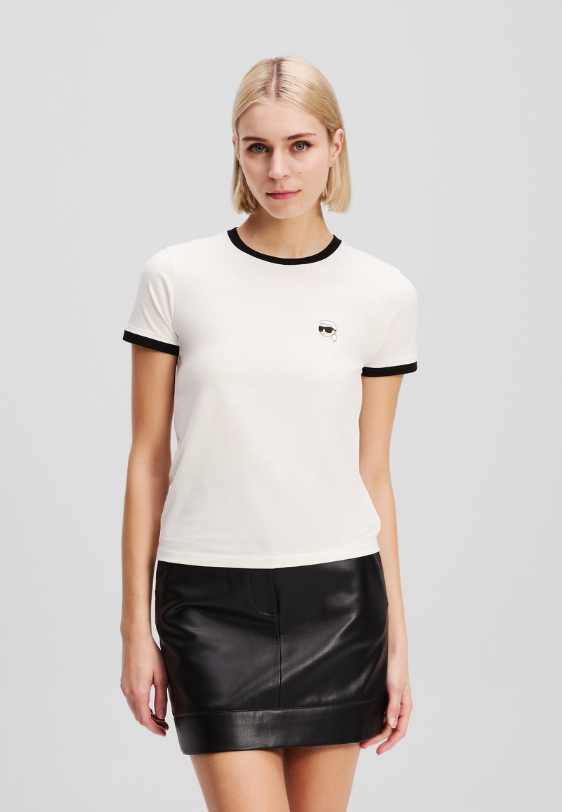 T-shirt blanc à manches courtes avec bord noir, présentant un petit graphique d'une personne portant des lunettes de soleil sur la poitrine. Matière en coton lisse.