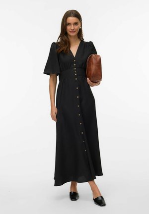 Kobieta stoi w czarnej, sięgającej do połowy łydki sukience zapinanej na guziki z bufkami na rękawach, trzyma brązową skórzaną kopertówkę, ma na nogach czarne szpilki, na tle białego, jednolitego tła.