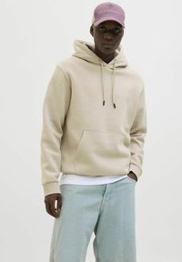 Beige Kapuzenpulli mit Kordelzügen, Fronttasche und gerippten Bündchen, kombiniert mit hellblauen weiten Jeans und einer lila Mütze.