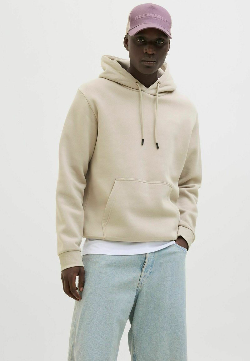 Beige Kapuzenpulli mit Kordelzügen, Fronttasche und gerippten Bündchen, kombiniert mit hellblauen weiten Jeans und einer lila Mütze.