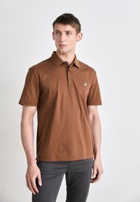 Barna pamut póló, galléros nyakkal, rövid ujjakkal és egy kis logóval a mellkason. Szürke nadrággal párosítva.