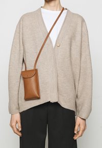 Beżowy sweter z dzianiny z zapięciem na guziki, zestawiony z brązową teksturowaną torbą na ramię. Luźny krój, długie rękawy i głęboki dekolt.