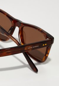 Ochelari de soare în culori de broască țestoasă cu lentile maro, design unghiular și un accent metalic lângă balama. Prezintă logo-ul "PRIVÉ REVAUX" pe ramă.