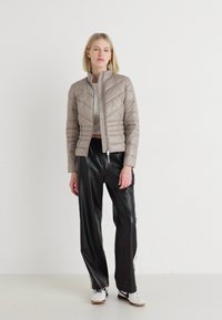 Lys beige quiltet jakke med høy krage, figursydd design og glidelås, kombinert med svarte vide bukser og hvite joggesko.