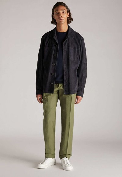 Dunkelblaues Wildlederjackett mit Knopfverschluss, zwei Fronttaschen, kombiniert mit olivgrünen Hosen und weißen Turnschuhen. Glatte Textur, lässige Passform.