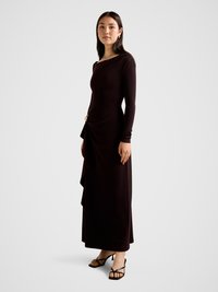 Long, fitted dark brown dress with long sleeves, asymmetric draping, and gold accent at the waist, paired with black strappy heels.