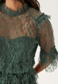 Abito in pizzo verde con scollatura alta e maniche trasparenti. Presenta dettagli arricciati e un design a strati, che mette in mostra bellissimi motivi in pizzo.