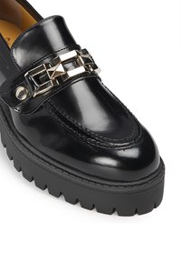 Mocasines de charol negro con suela gruesa negra, que presentan un detalle de cadena geométrica metálica en el empeine. Perfil plano y textura suave.