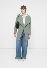 Zöld gyapjú keverék kabát széles gallérral, világoskék, bő szárú farmerral, fehér inggel és egy kis fekete crossbody táskával párosítva.