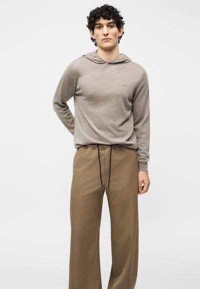 Jeune homme aux cheveux bouclés portant un sweat à capuche marron clair et un pantalon ample beige avec un cordon noir, debout devant un fond uni.