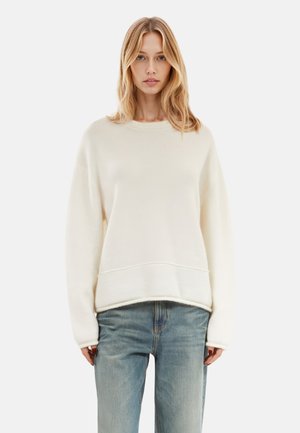 Cremefarbener Pullover mit Rundhalsausschnitt und langen Ärmeln. Weiche Textur und lockere Passform, mit einer Nahtdetail am Saum. Kombiniert mit hellblauen Jeans.