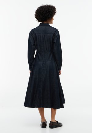 Kvinde med krøllet hår står med ryggen mod og bærer en mørk langærmet denimkjole og sorte loafers på en hvid baggrund.