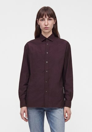 Femme portant une chemise boutonnée manches longues violet foncé et un jean bleu clair, debout devant un fond blanc uni.