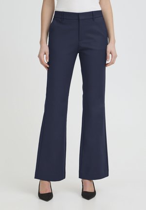 Ženska v mornarsko modrih širokih hlačah po meri in črnih čevljih z ostrim prstom, stoječa pred enobarvnim belim ozadjem.