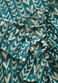 Tissu bleu-vert avec un motif floral et de feuillage en blanc. Présente une bordure froncée et des attaches décoratives, texturé avec une finition douce et légère.
