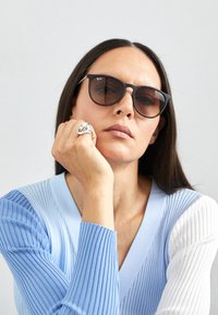 Black sunglasses with a slight cat-eye shape, worn with a light blue sweater featuring ribbed texture and white accents, displaying a silver ring.