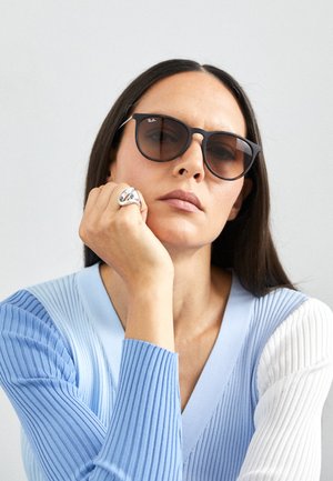 Zwarte sunglasses met een lichte cat-eye vorm, gedragen met een lichtblauwe trui met ribbeltextuur en witte accenten, en een zilveren ring zichtbaar.