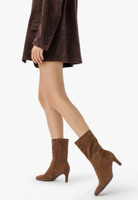 Kvinna som bär bruna mocka ankelboots med kitten heels och en mörkbrun mockakappa, som går mot en vit bakgrund.