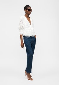 Nő fekete napszemüvegben, fehér csipkés blúzban, bő ujjú, kék farmernadrágban és barna magassarkú szandálban áll, kezét zsebre téve.