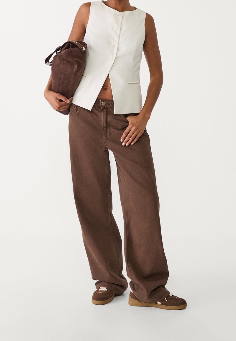 Veste blanche sans manches à cinq boutons, associée à un pantalon large marron et des baskets marron. Un sac à bandoulière en cuir marron est tenu.