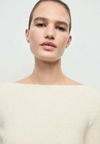 Giovane donna con capelli pettinati all'indietro che indossa un maglione a coste di colore bianco sporco, rivolta in avanti con espressione neutra su uno sfondo chiaro e uniforma.