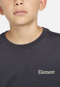 Camiseta de algodón gris oscuro con cuello redondo y mangas cortas. Presenta un pequeño logo crema bordado con la palabra "Element".