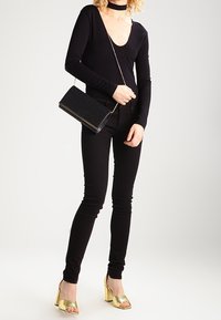 Svart långärmad topp med djup V-ringning, svarta skinny jeans, guld blockklacksandaler och en svart texturerad clutchväska med silverkedja.