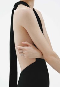 Prenda sin mangas negra con un escote profundo en la espalda, detalle de cuello atado y una silueta ajustada. Textura suave y un corte lateral. Anillo plateado en la mano.