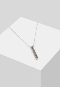 Collier pendentif en argent avec une barre rectangulaire texturée suspendue à une fine chaîne, présenté sur une surface blanche inclinée.