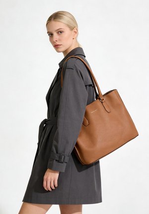 Femme aux cheveux blonds portant un manteau gris foncé ceinturé et un grand sac cabas en cuir marron sur l'épaule.