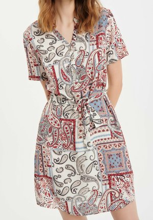 Vrouw draagt een korte mouw jurk met ceintuur, versierd met rood, blauw en zwart paisley- en patchworkpatronen op een witte achtergrond.