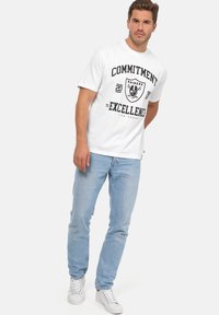 Camiseta blanca de algodón con texto gráfico negro "COMPROMISO CON LA EXCELENCIA" y logo. Combinada con jeans de denim azul claro y zapatillas blancas.
