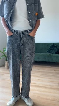 Denim kombinezon, ki vključuje sivo majico z kislim pranjem in hlače z visokim pasom z teksturirano površino. Bele superge dopolnjujejo videz.