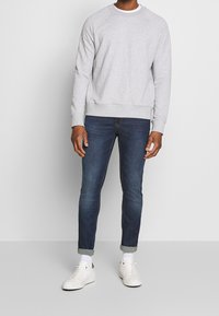 Šedá mikina s kulatým výstřihem a raglánovými rukávy, v kombinaci s tmavě modrými slim fit džíny. Bílé tenisky s ohrnutými manžetami, ukazující bílé ponožky.