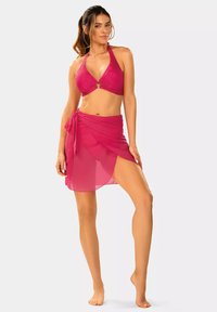 Jaskrawo różowy bikini z fakturowanym topem na halter i okrągłym akcentem. W komplecie przezroczysta spódnica w stylu owijanym. Gładki materiał układa się elegancko.