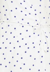Tissu blanc avec des pois bleus régulièrement espacés, présentant un détail à nouer à la taille et une texture douce et lisse.