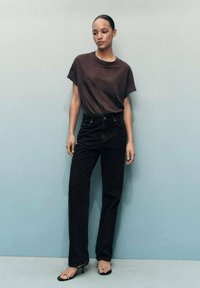 Black high-waisted jeans paired with a dark brown short-sleeve T-shirt. Model stands against a soft blue background.