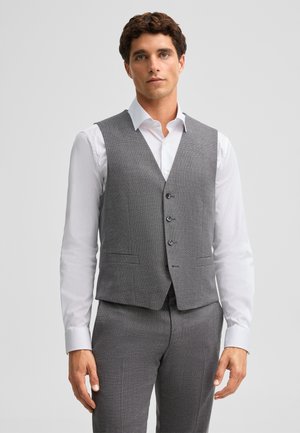 Homme aux cheveux courts et bouclés portant un gilet de costume gris et un pantalon assorti sur une chemise blanche, debout devant un fond uni.