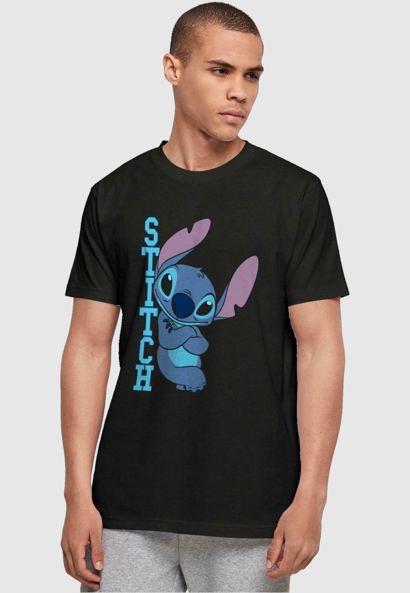 Zwarte t-shirt met korte mouwen met een grafisch ontwerp van een blauwe figuur met grote paarse oren en het woord "STITCH" in blauw ernaast.