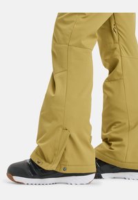 Pantaloni da neve gialli con vestibilità rilassata, dotati di zip ai polsini e orli rinforzati, abbinati a stivali da neve neri e bianchi.