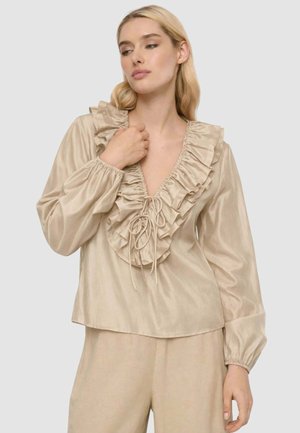 Donna bionda che indossa una blusa di raso beige con maniche lunghe e scollatura a V con volant, legata con un laccetto, abbinata a pantaloni beige.
