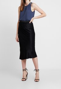 Top senza maniche blu navy con scollo arricciato, abbinato a una gonna midi nera aderente in satin e sandali con tacco a strappi neri.